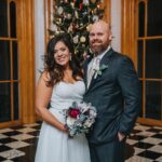 Elopement Wedding A smiling bride in a white dress holds a bouquet of flowers while standing next to a groom in a dark suit and tie. They are in front of a decorated Christmas tree and wooden framed windows in a venue with checkered black and white floors. Elopements Inc